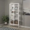 ויטרינת מתכת לבנה עם דלתות זכוכית מצולעת דגם 205, בסלון עם ספרים, רדיו וסלסלות קש, ידיות זהב