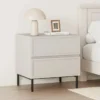 שידת לילה בגוון סהרה עם שתי מגירות מסגור ורגלי מתכת שחורות לצד מיטה, מנורה ומסגרת תמונה על המשטח - הדמיית חדר